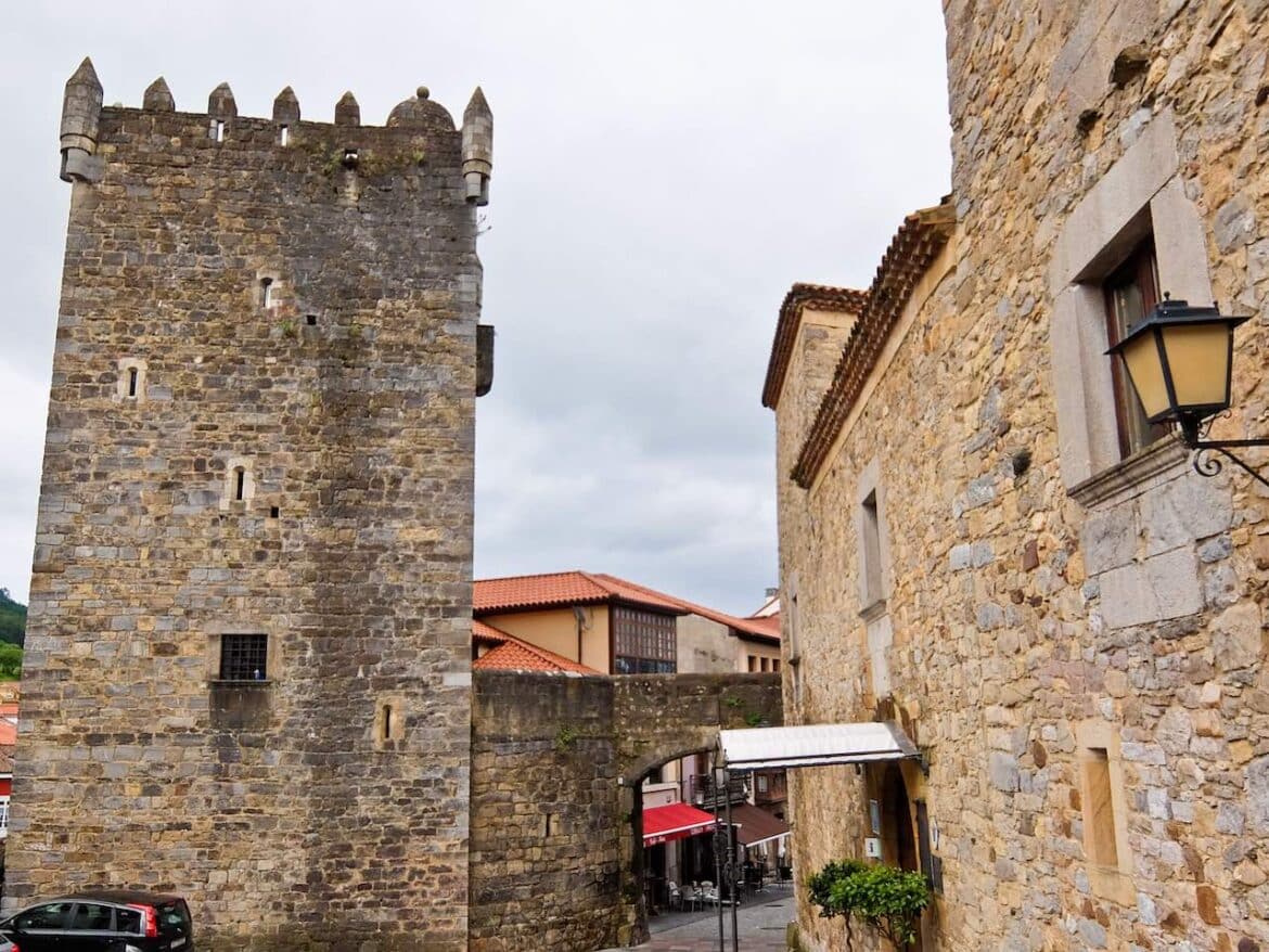 Stone medieval tower with crenellations beside a stone building on a narrow street; outdoor café tables visible in the background beneath arches.