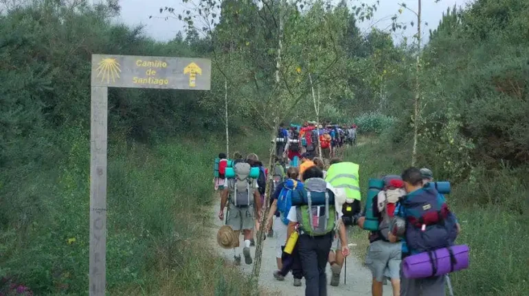 Hikers with large backpacks walk along a dirt trail beside a Camino de Santiago sign at the start of the path.