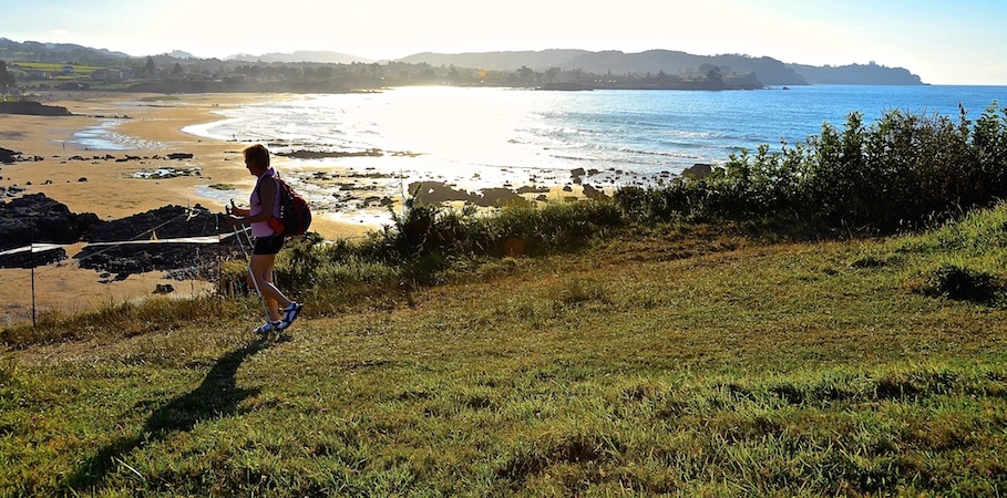 Hiker with backpack walking along a grassy coastal overlook toward a rocky beach and the sea at sunset.