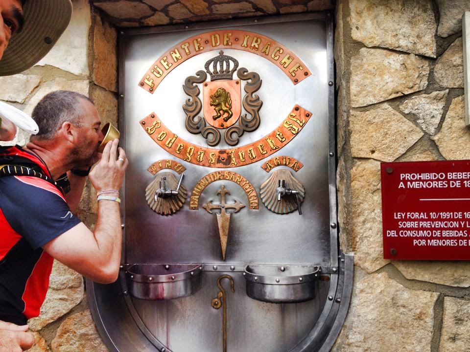 Man drinking from a historic metal fountain set in a stone wall, with an ornate crest above two spouts and a red informational plaque nearby.