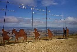 Row of camel-shaped sculptures with riders on a pebbly beach, under a blue sky with the sea in the background.
