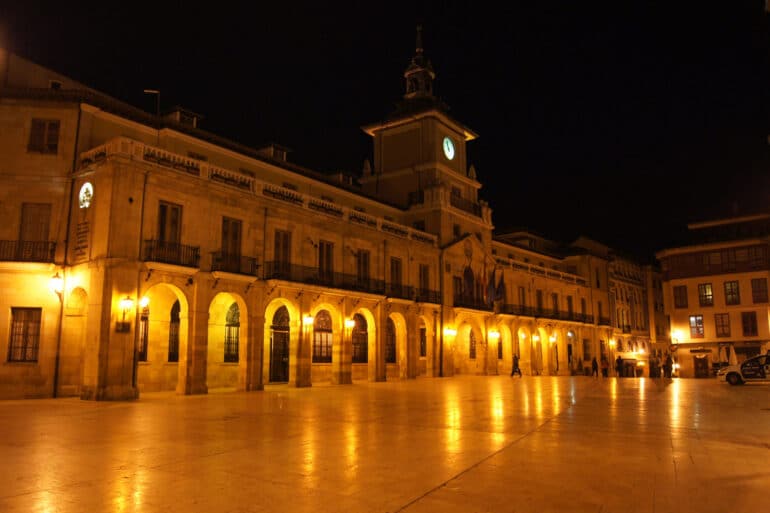 Nighttime plaza with a illuminated arched colonnade and clock tower building, warm yellow lights reflecting on the wet-looking ground