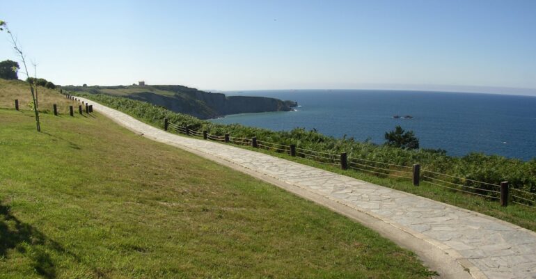 Coastal park with a stone path winding along grassy cliffs and a rope fence, overlooking the blue sea and horizon.