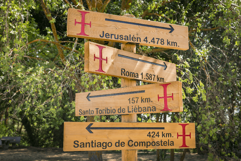 Wooden signpost with arrows listing distances to Jerusalem, Rome, Santo Toribio de Liébana, and Santiago de Compostela in a forest setting.