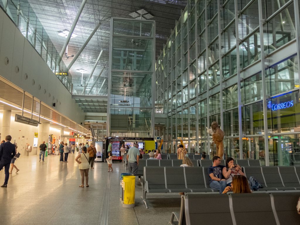Busy airport terminal with glass walls, metal framework, and rows of seats as travelers walk and wait.