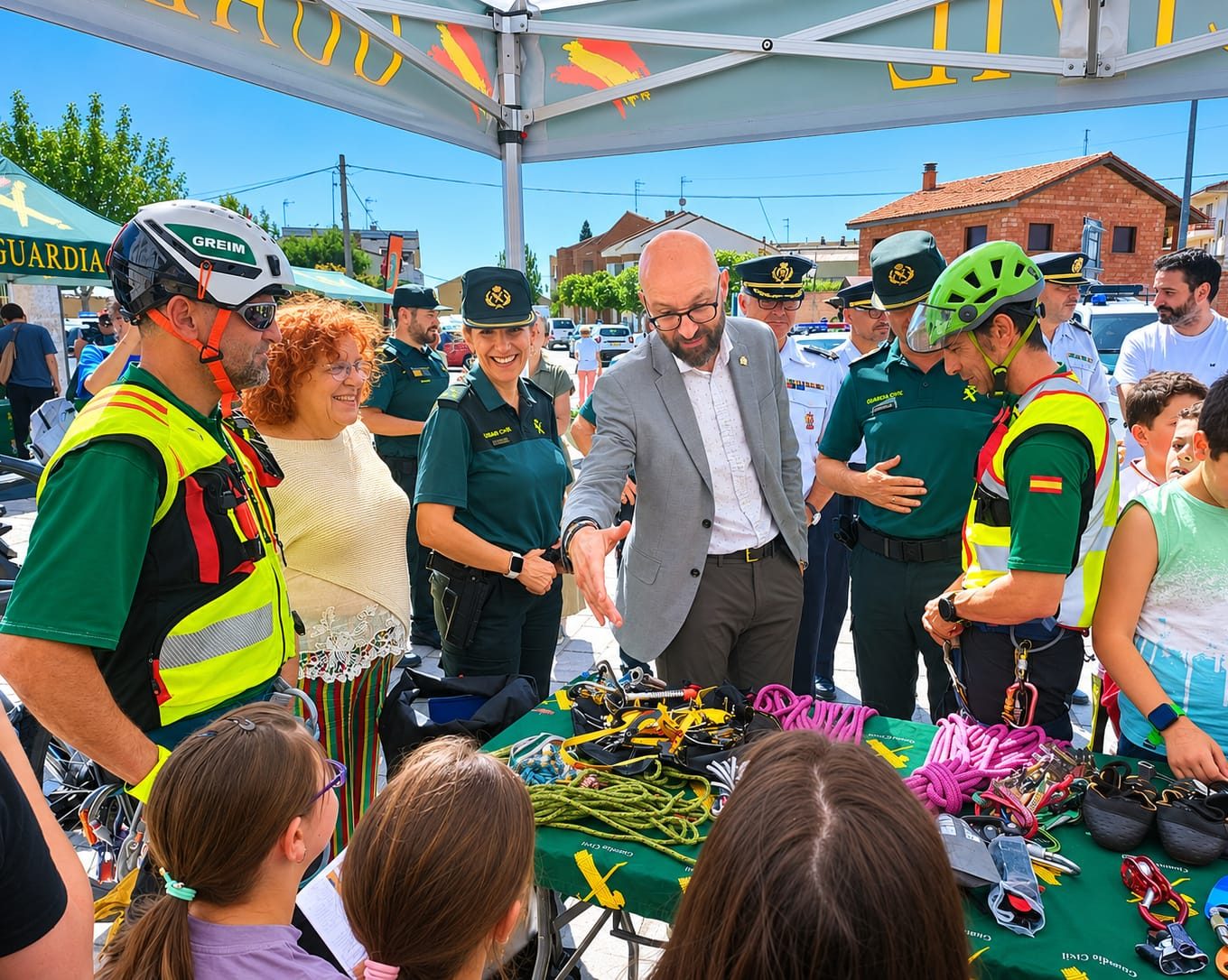 Public safety demonstration: rescue workers and civilians gather around a table of climbing gear (harnesses, ropes) while a man in a suit speaks.