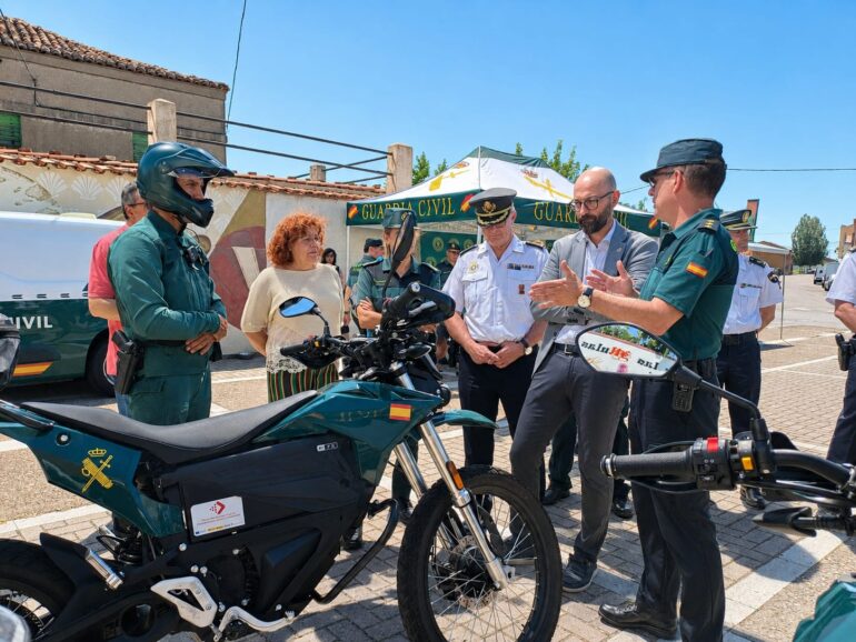 Group of Guardia Civil officers and civilians standing near two teal motorcycles at an outdoor event.