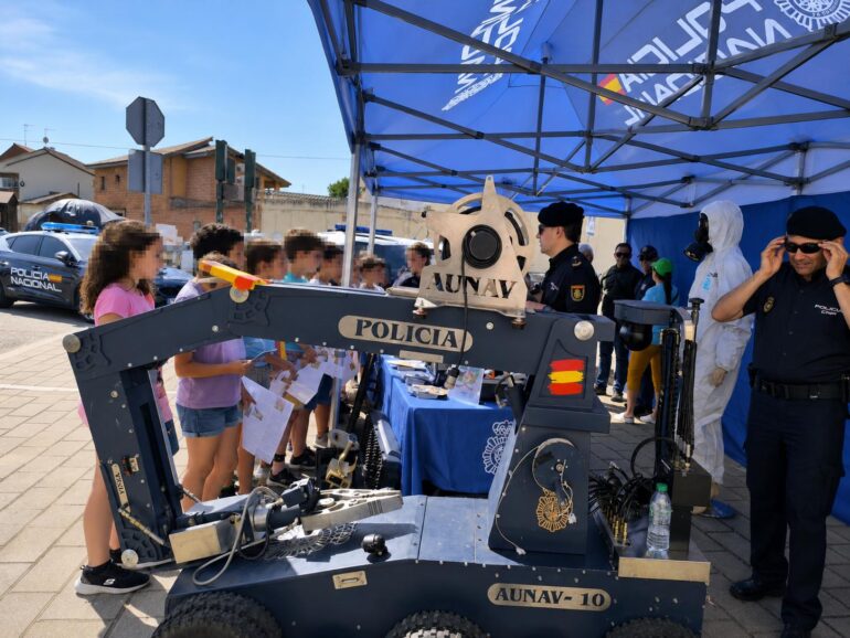 Public safety event: children watch a large robotic arm labeled POLICIA AUNAV-10 exhibited by police under a blue tent.