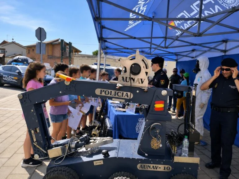 Veranstaltung zur öffentlichen Sicherheit: Kinder beobachten einen großen Roboterarm mit der Aufschrift POLICIA AUNAV-10, der von der Polizei unter einem blauen Zelt ausgestellt wird.