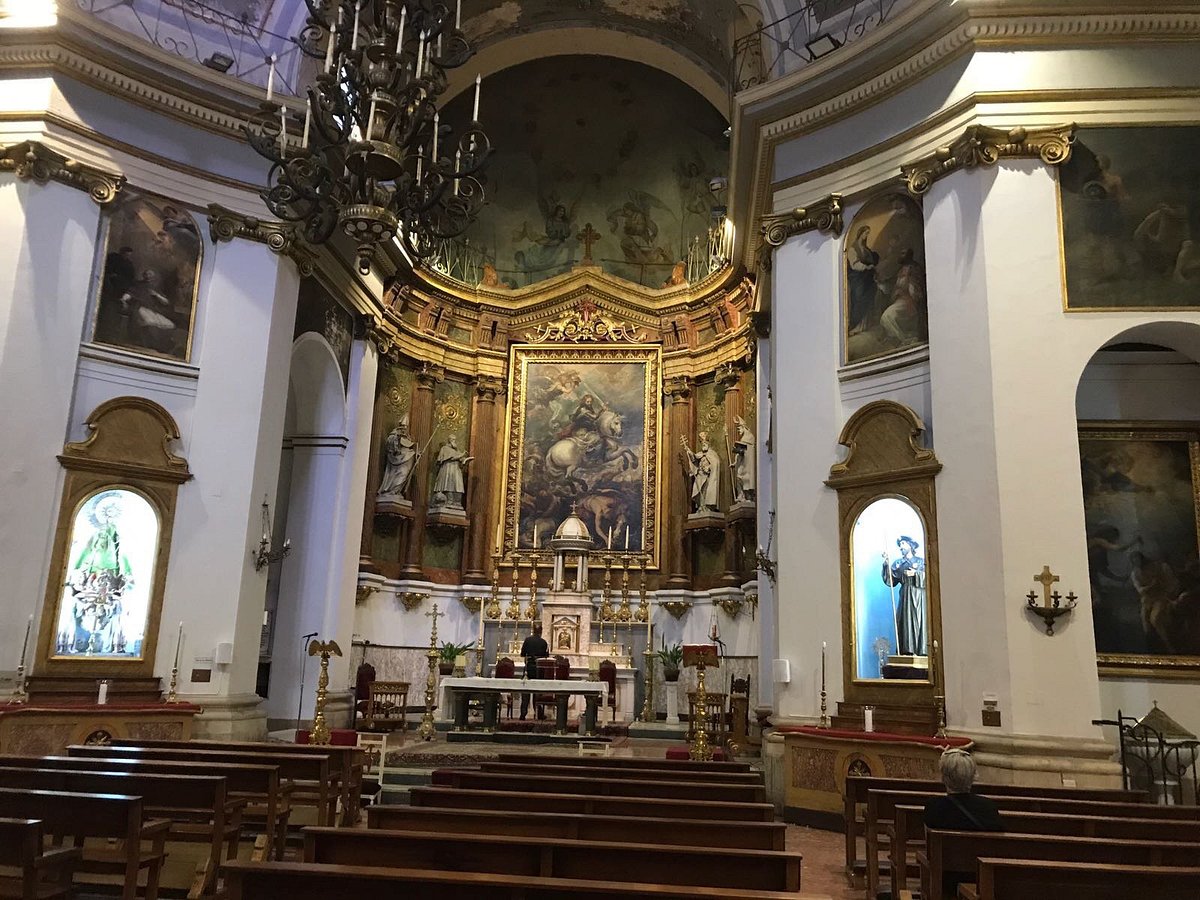 Interior of a ornate church sanctuary featuring a central altar with a large framed painting, statues, and gold decor along curved chancel walls under a chandelier.