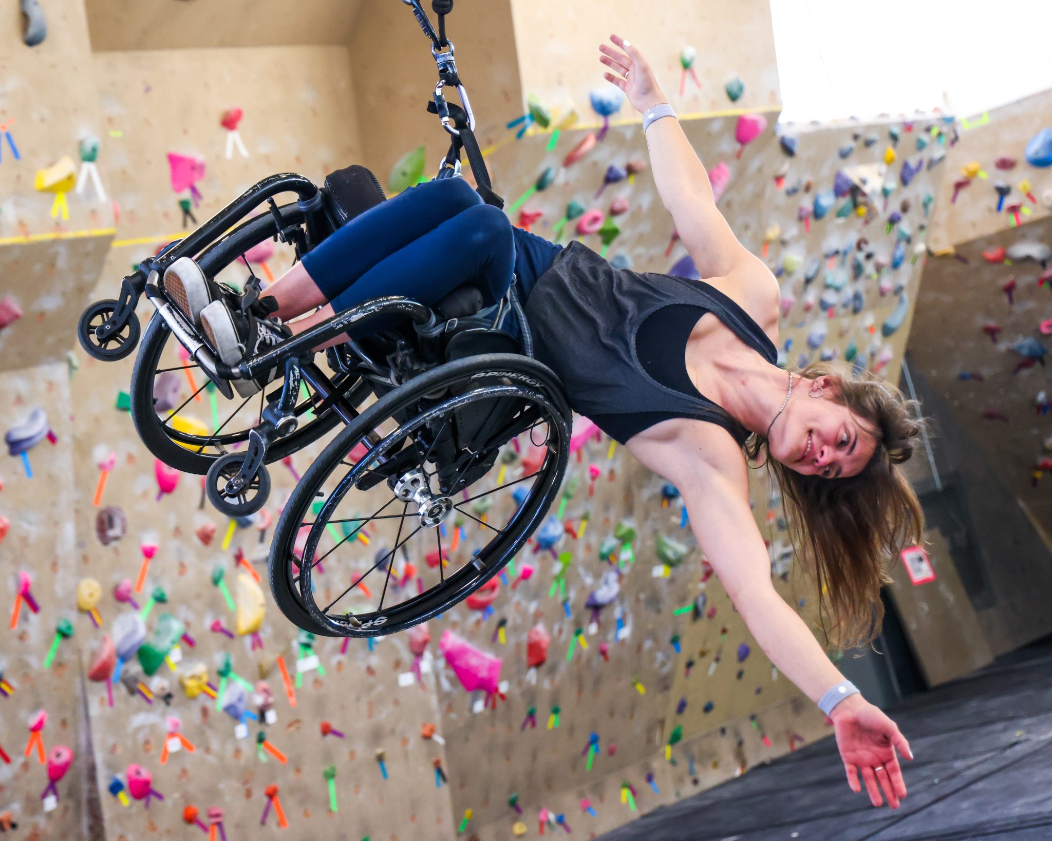 Wheelchair user climbing an indoor rock wall, suspended with harnesses and reaching toward the wall with outstretched arm.