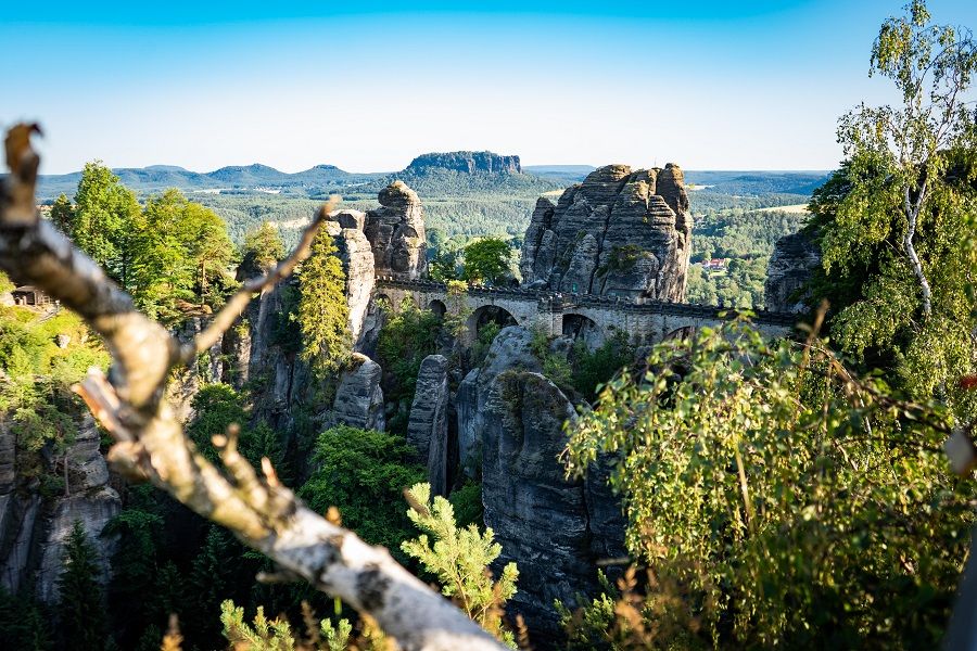Stone arch bridge spans a deep, rocky canyon, surrounded by green trees under a bright blue sky on a sunny day