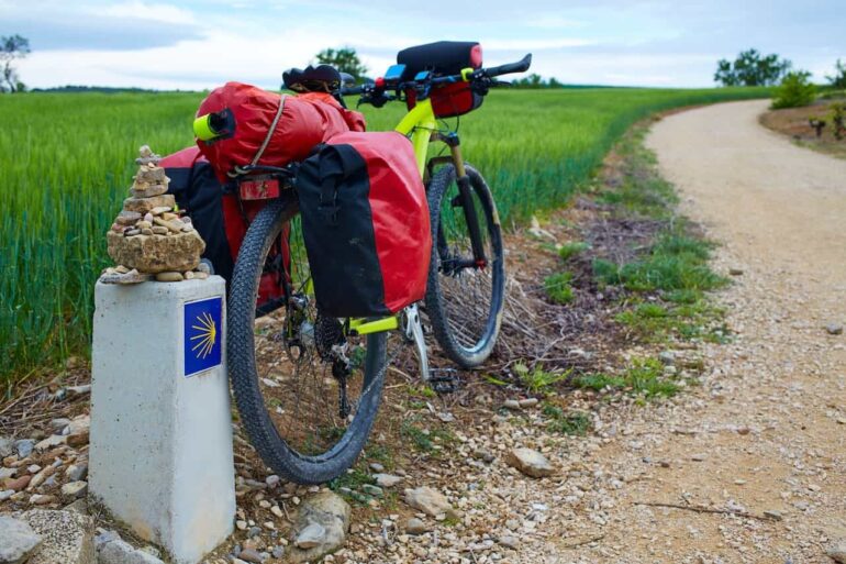 Touring bicycle loaded with red panniers parked on a gravel path beside a Camino marker in a rural field setting (sacred shell symbol).
