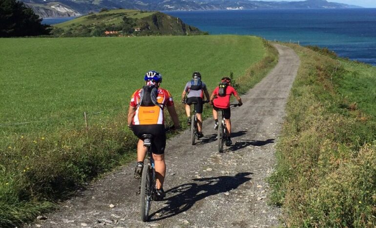 Three cyclists ride along a gravel coastal trail with green fields and blue sea on either side.