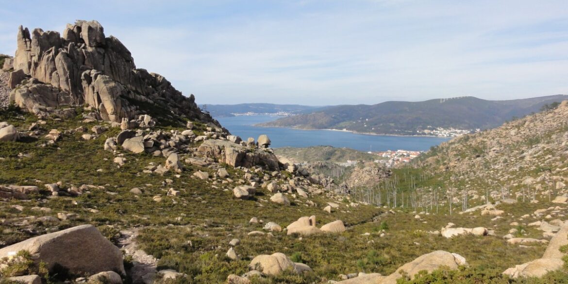 Rugged rocky hillside with large boulders, sloping toward a blue bay and distant town beyond.