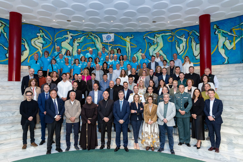 Large group photo of men and women posed on marble stairs inside a mural-adorned hall, some in formal attire and uniforms, smiling at the camera.