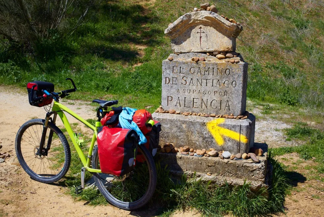 Bright lime-green touring bike with red panniers parked beside a stone milestone on the Camino de Santiago path, yellow arrow painted on the base.