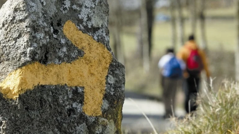 Rock in foreground with a yellow trail marker painted on it; hikers with backpacks blur in the background on a path.