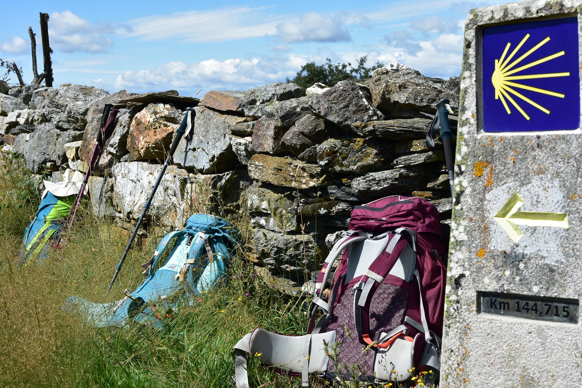Hiking gear—backpacks and trekking poles—resting against a rough dry stone wall under a blue sky, with a Camino scallop shell marker nearby.