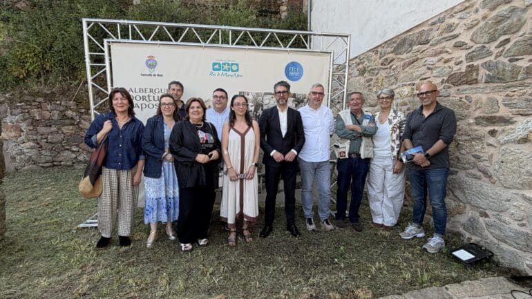 Twelve adults pose for a group photo outdoors in front of a banner with logos, with a stone wall and greenery in the background.