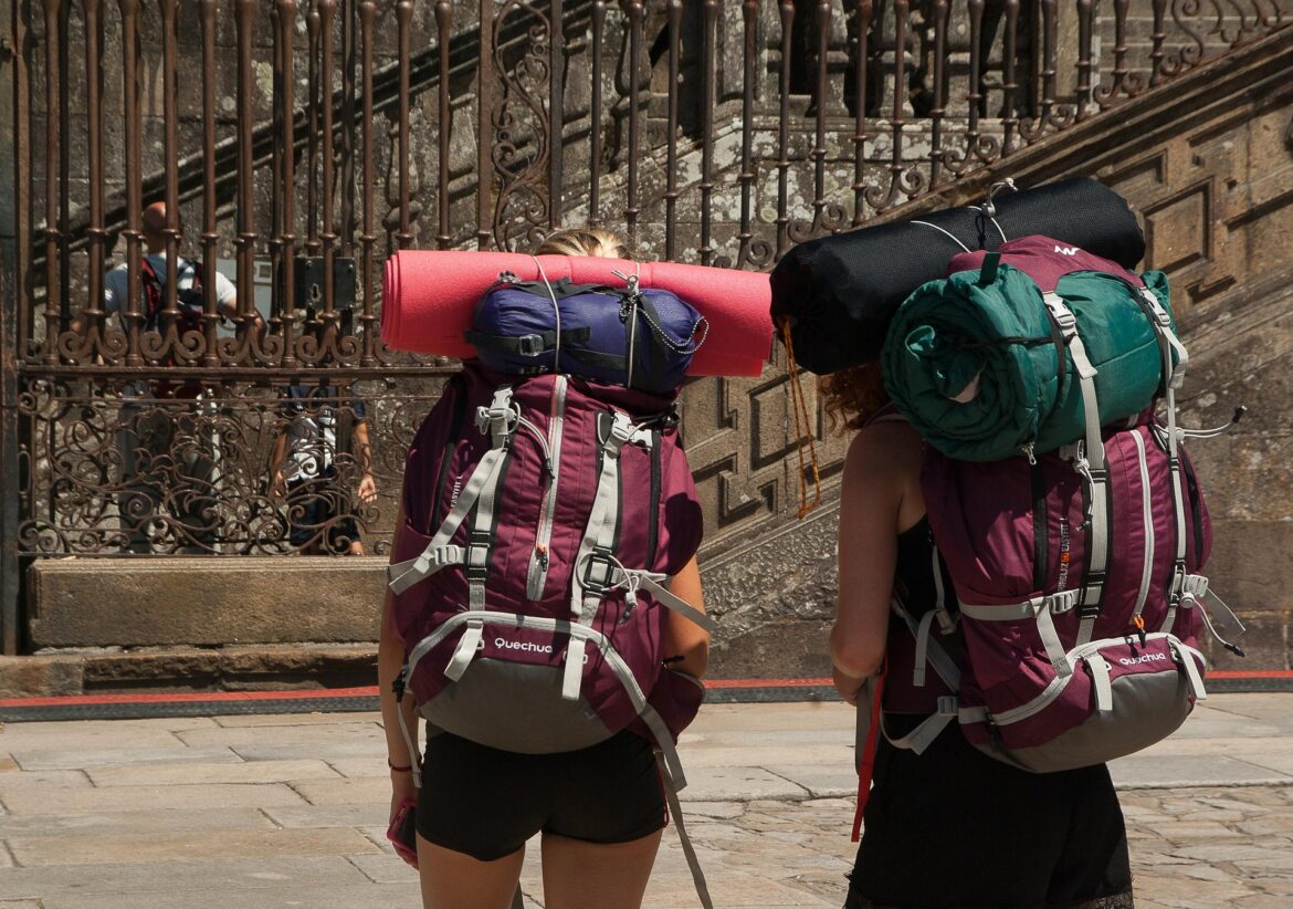 Two hikers with large backpacks and a rolled-up mat walk past an ornate iron railing, seen from behind.