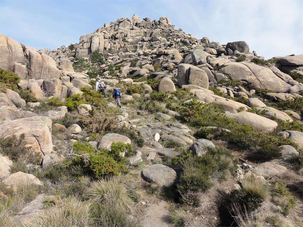 Two hikers with backpacks climb a rocky, boulder-strewn trail toward a hill of large granite rocks in a desert landscape.