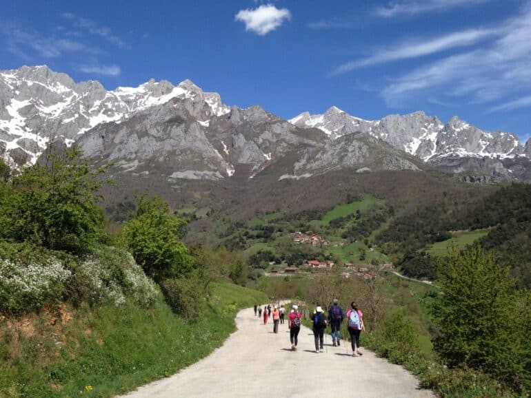 Group of hikers walking a dirt road through a green valley toward a village with snow-capped mountains under a blue sky