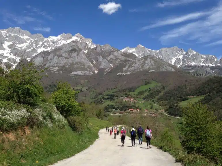 Group of hikers walking a dirt road through green valley toward village with snow-capped mountains under blue sky