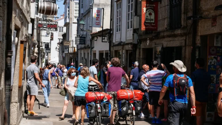 Busy narrow street in a historic town with many pedestrians, some wearing masks, and bicycles with large red panniers in the foreground.