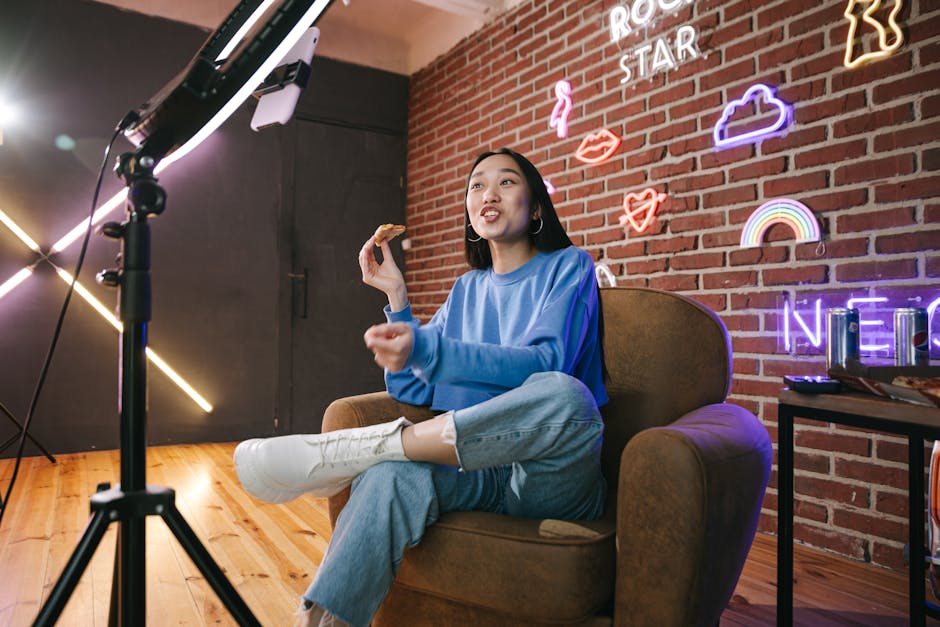 Video creator seated in a brown armchair in a neon-lit studio, speaking and gesturing during a livestream setup with a ring light nearby.