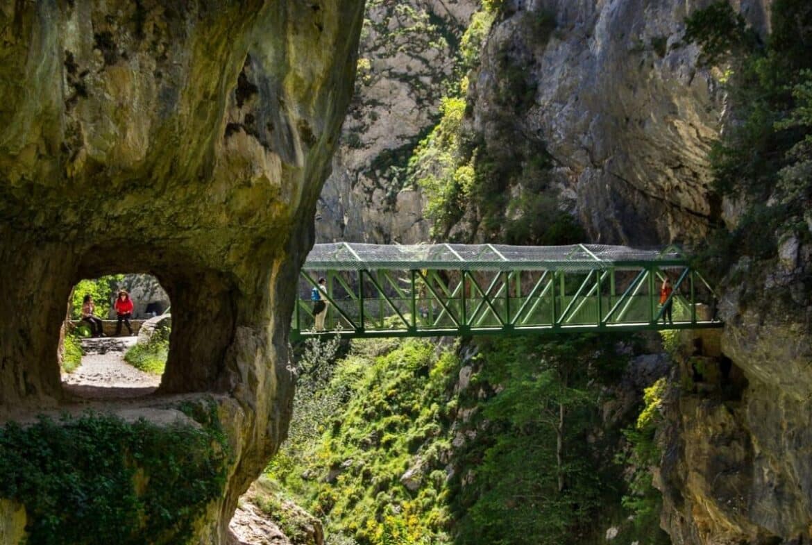 Green metal footbridge spans a deep rocky gorge, with hikers crossing between steep cliffs.