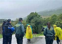 Group of rescue workers in rain gear standing on a wet road in a misty, hilly landscape.