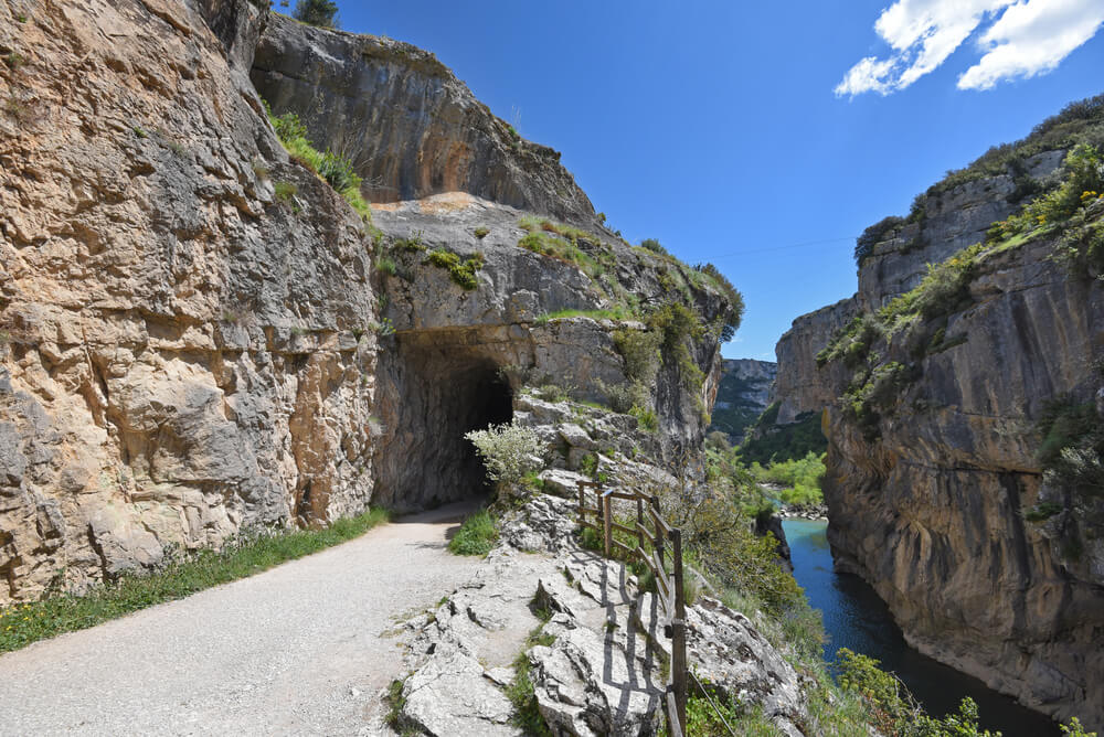 Trail along a rocky cliff with a dark tunnel entrance on the left, a wooden railing, and a blue river visible in the canyon below under a clear sky.