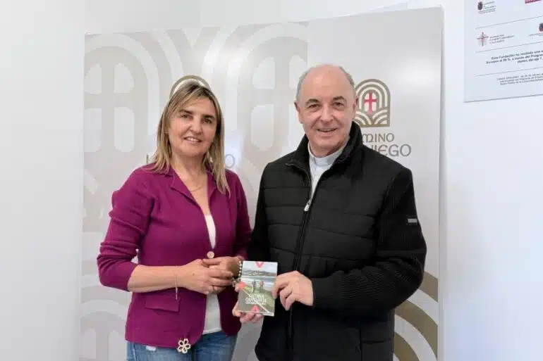 Dois adultos estão dentro de casa, sorrindo, enquanto a mulher de blazer magenta entrega um pequeno livreto ao homem de jaqueta preta em frente a um cenário de marca.
