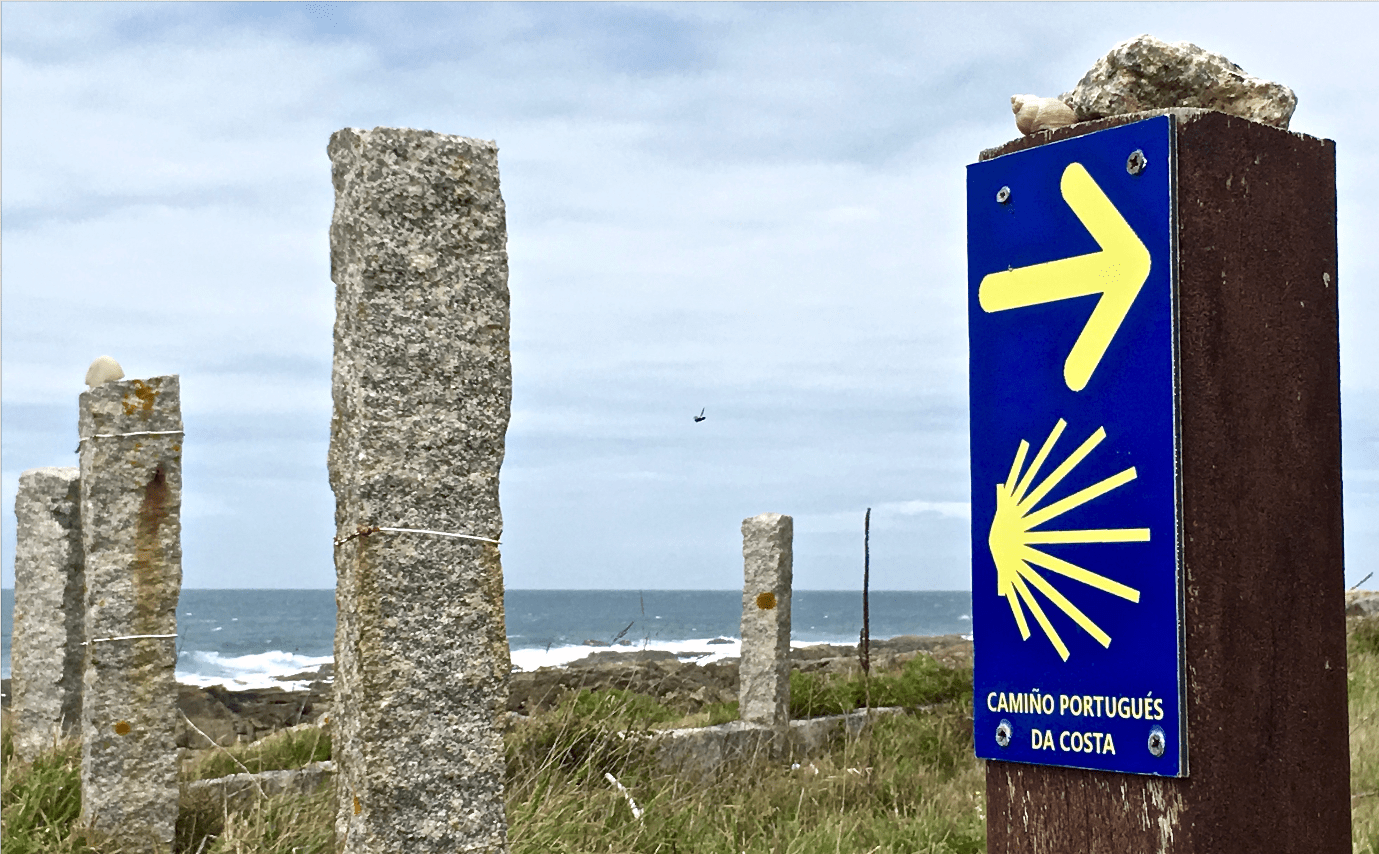 Blue Camino Portuguese Coastal sign with a yellow arrow and shell symbol next to weathered stone posts by the sea coast.