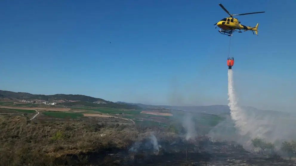 Yellow helicopter with a water bucket dumping water onto a rural brush fire, over a dry, smoky landscape.