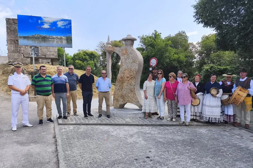Group of people posing in front of a large stone sculpture in a park; mix of casual clothes and traditional dresses, a uniformed man on the left.