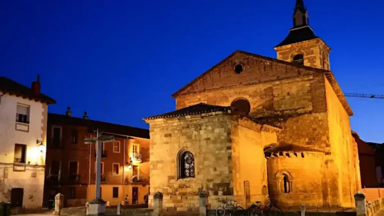 Stone church at night illuminated by warm lights, with surrounding houses and a blue evening sky nearby.