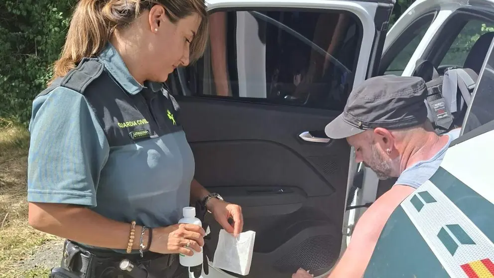 Female Guardia Civil officer hands sanitizer and a wipe to a man by an open car door near a police vehicle.