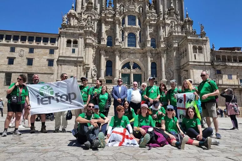 Group of about 20 people in matching green shirts pose for a photo in a sunny square, in front of an elaborate stone cathedral, with some holding a white banner with a logo.