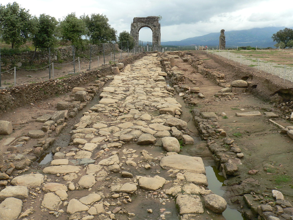 Ancient stone road with large cobbles leading to a ruined arch and distant hills under a cloudy sky, fenced archaeological site on both sides.