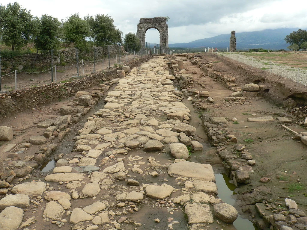 Ancient stone road with large cobbles leading to ruined arch and distant hills under a cloudy sky, fenced archaeological site on both sides.