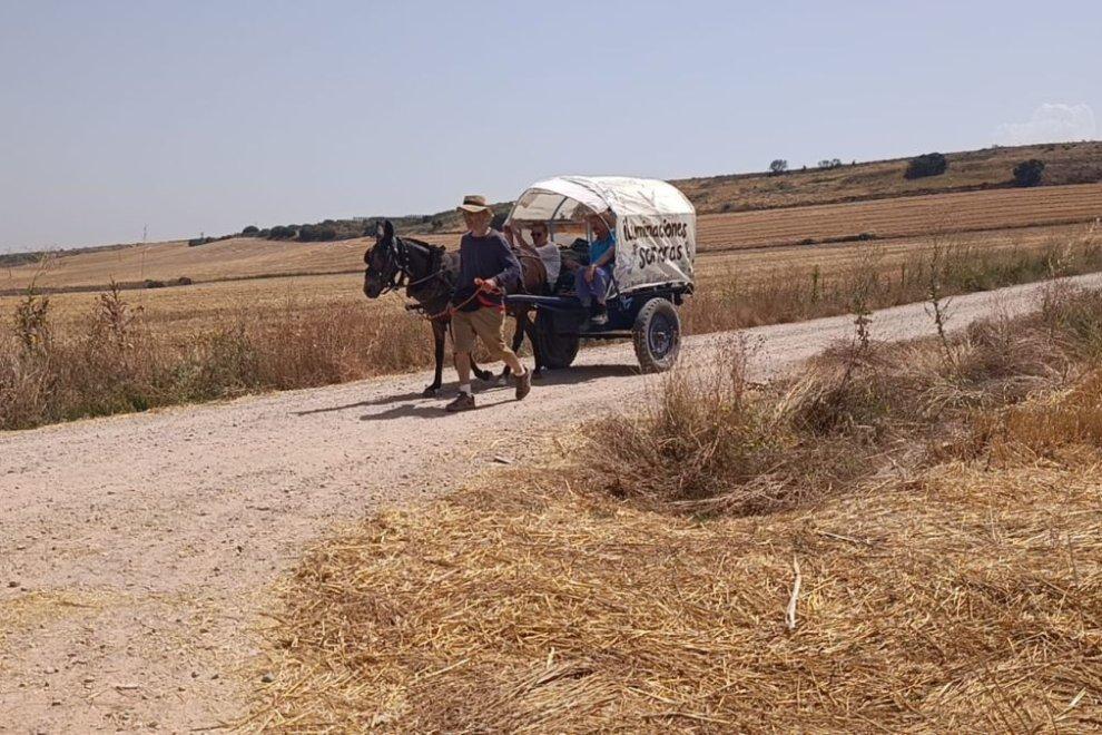 Man leading a horse-drawn wagon on a rural dirt road; covered wagon reads 'Iluminaciones Solares' with people riding inside.