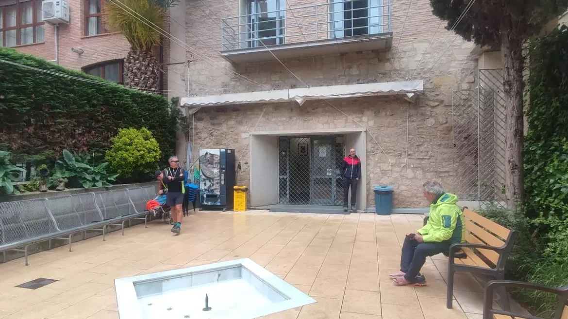 Open-air courtyard in front of a stone building with benches, a small square fountain, and several people: a man with a backpack walking, a person in a bright jacket sitting on a bench, and another person near the entrance.