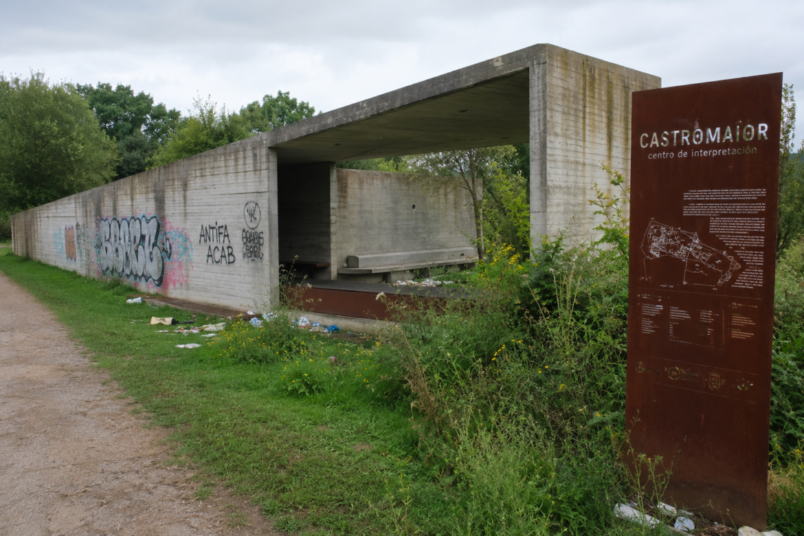 Concrete open-air pavilion with graffiti along a dirt path; a rust-colored interpretive sign stands to the right, reading 'CASTROMAIOR centro de interpretación'.