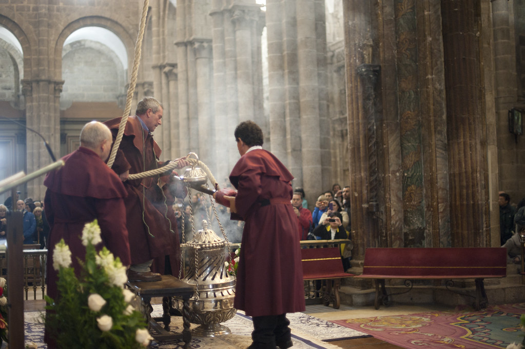 Monks in burgundy robes perform a religious blessing with a silver incense censer inside a stone church, while onlookers watch from the background.