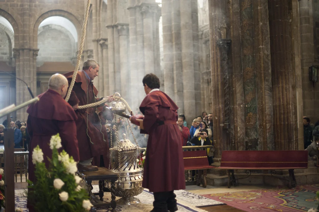 Mönche in burgunderroten Gewändern vollziehen in einer Steinkirche einen religiösen Segen mit einem silbernen Räuchergefäß, während Zuschauer im Hintergrund zuschauen.