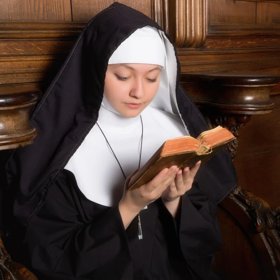 Nun in a black habit reading a worn Bible while seated on a wooden bench or pew.