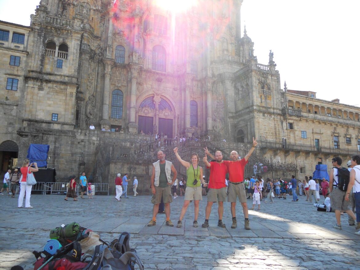 Four people pose with arms raised in front of a grand ornate cathedral; camping gear and backpacks on the stone plaza in the foreground.