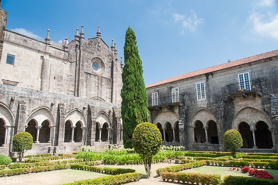 Stone cloister with arched walkways surrounding a formal garden and central topiary, under a bright blue sky